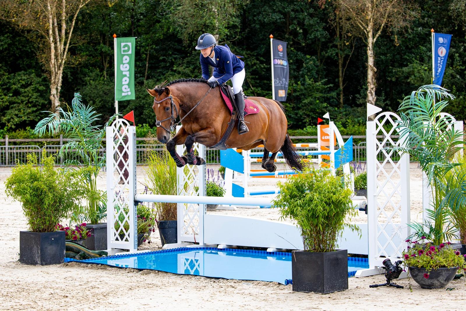 Lynn en een bruin paard springen over een blauw-witte hindernis tijdens een concours.
